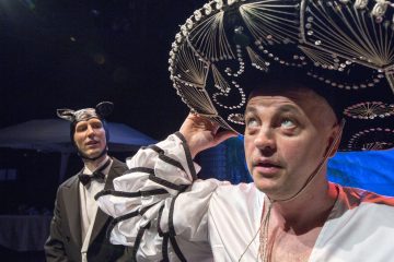 Lavalla kaksi pukumiestä; toisella on suuri, koristeltu sombrero ja valkoinen paita, kun taas toinen on pukeutunut smokkiin, jossa on kissankorvat ja rusetti. Molemmat katsovat mietteliäästi kaukaisuuteen teatterin valaistuksessa.