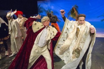 Värikkäisiin, taidokkaisiin kuninkaallisiin pukuihin pukeutuneet näyttelijät esittävät dramaattisen kohtauksen lavalla, jonka taustalla on trooppisia kulisseja ja palmuja. Yksi näyttelijä näyttää lyövän toista, kun muut katselevat taustalla.