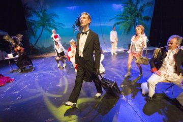 Smokkiin pukeutunut mies kävelee dramaattisesti lavan poikki, kun muut eklektisiin pukuihin pukeutuneet poseeraavat hänen ympärillään. Taustalla on palmuja ja sininen taivas, mikä viittaa trooppiseen ympäristöön. Kohtauksessa on teatraalinen tunnelma.