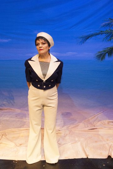 Merimiesasuun ja -hattuun pukeutunut henkilö seisoo lavalla, jonka taustalla on sininen meri, hiekka ja palmu.