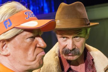 Kaksi pukumiestä: toisella on oranssi visiiri ja hän nyrpistelee otsaansa, kun taas toinen, ruskeaan hattuun ja turkistakkiin pukeutunut mies katsoo häntä vakavalla ilmeellä ja suurilla viiksillä. Taustalla on tumma sininen valaistus.