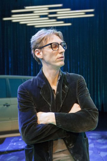 Lyhyet, vaaleanruskeat hiukset ja silmälasit omaava henkilö seisoo kädet ristissä, yllään tumma bleiseri. Taustalla on auto ja vaakasuoria valoja sinistä verhoa vasten.