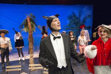 Smokkiin ja kissankorviin pukeutunut mies seisoo näyttämön keskellä kädet kohotettuna, ympärillä on värikkäisiin, omituisiin pukuihin pukeutuneita ihmisiä, kuten noita ja punapukuinen nainen, ja taustalla on trooppinen ranta.