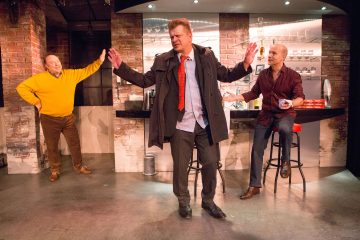 Kolme miestä on modernilla lavasteella, joka muistuttaa baaria. Yksi keltaiseen puseroon pukeutunut mies seisoo hymyillen käsi lantiollaan, kun taas takkiin pukeutunut, epäsiisti mies elehtii laajasti. Kolmas mies istuu ja katselee kuppi kädessään.