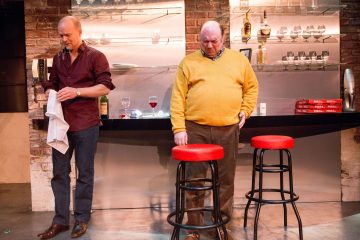 Kaksi miestä seisoo modernissa keittiössä, jossa on tiiliseinät; toinen pyyhkii lasia pyyhkeellä, kun taas toinen, keltainen villapaita yllään, katsoo alaspäin. Kaksi punaista baarijakkaraa ja viinilasit ovat tiskillä heidän takanaan.