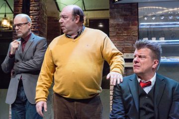 Lavalla on kolme miestä: yksi silmälasipäinen pukumies katsoo mietteliäästi, yksi keltaiseen puseroon pukeutunut mies seisoo keskellä, ja yksi pukumies, jolla on punainen rusetti, istuu ja katsoo sivulle. Lavastus näyttää olevan sisätiloissa.