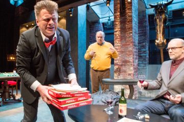 Pizzalaatikoita kantava pukumies näyttää ahdistuneelta. Hänen takanaan keltaiseen puseroon pukeutunut mies pitelee juomaa, ja toinen silmälasipäinen mies istuu viinilasin kanssa. Huone on hämärästi valaistu, ja siinä on moderni sisustus ja asennettu hirvenpää.