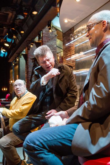 Kolme miestä istuu ja vuorovaikuttaa teatteritilassa. Yksi pukumies elehtii kädellään ilmeikkäästi, kun taas kaksi muuta kuuntelevat tarkkaavaisesti. Taustalla näkyy hyllyjä, joissa on laseja ja ylävaloja.