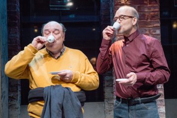 Kaksi vanhempaa miestä seisoo vierekkäin siemaillen pienistä valkoisista kupeista. Toisella on keltainen pusero ja takki kädessä, toisella viininpunainen paita ja silmälasit. He ovat tiiliseinän edessä, mahdollisesti rennossa seurapiiritapahtumassa.