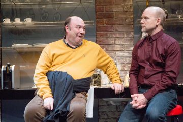 Kaksi miestä istuu keittiössä; toisella on keltainen pusero ja hän hymyilee puhuessaan, toisella on ruskeankeltainen paita ja hän kuuntelee vakavana. Taustalla näkyy hyllyjä, joissa on kuppeja ja laseja, sekä tiiliseinä.