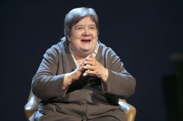 Vanhempi nainen, jolla on lyhyet harmaat hiukset, istuu tuolilla ja hymyilee lämpimästi kädet yhteen lyötyinä. Hänellä on yllään harmaa asu, joka on asetettu tummaa, yksinkertaista taustaa vasten.