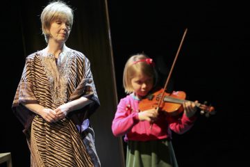 Aikuinen seisoo ja katselee, kun vaaleanpunaiseen neuletakkiin pukeutunut nuori tyttö soittaa viulua. Tausta on tumma, ja molemmat ovat pehmeästi valaistuja, mikä viittaa esitykseen tai musiikkituntiin.