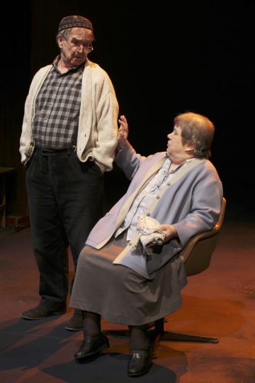 Vanhempi mies, jolla on neuletakki, seisoo tuolilla istuvan vanhemman naisen vieressä ja elehtii, kun nainen puhuu hänelle hämärästi valaistulla näyttämöllä. Molemmat ovat pukeutuneet rentoihin vaatteisiin, mikä viittaa dramaattiseen teatterikohtaukseen.