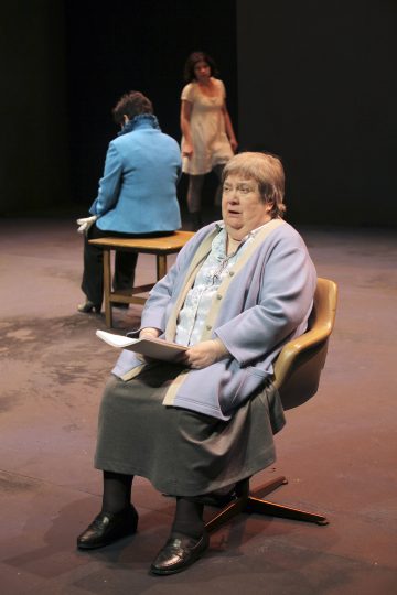 Lavalla on kolme naista. Yksi, siniseen villatakkiin pukeutunut, istuu etualalla pitelemässä papereita. Toinen istuu penkillä selin, ja kolmas seisoo taustalla valkoisessa mekossa. Näyttämö on lavastettu minimaalisesti.