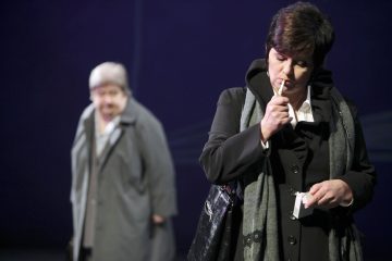 Tummiin takkeihin pukeutunut nainen sytyttää savukkeen pitelemällä aski kädessään, ja toinen harmaaseen takkiin pukeutunut vanhempi nainen seisoo epäselvästi taustalla tummaa taustaa vasten.