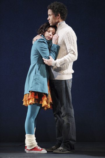 Siniseen takkiin ja värikkääseen hameeseen pukeutunut nuori nainen halaa valkoiseen villapaitaan ja farkkuihin pukeutunutta nuorta miestä. Nainen painaa päänsä miehen rintaan, kun mies pitää häntä hellästi sylissään, molemmat seisovat yksinkertaista tummaa taustaa vasten.