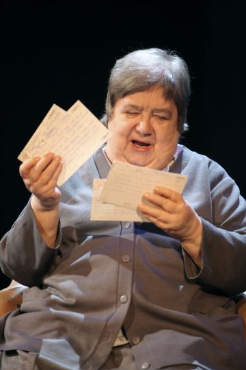 Vanhempi nainen, jolla on lyhyet harmaat hiukset ja harmaa napitettu asu, istuu ja tutkii mietteliäästi käsinkirjoitettuja kirjeitä tai kortteja molemmissa käsissään tummaa taustaa vasten.