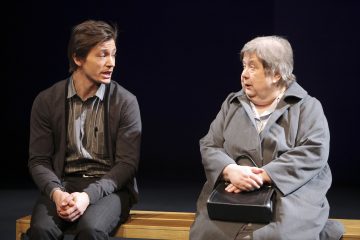 Nuori mies ja vanhempi nainen istuvat puupenkillä keskustellen. Mies elehtii käsillään, kun taas nainen kuuntelee tarkkaavaisesti pitäen sylissään mustaa käsilaukkua. Molemmat vaikuttavat vakavilta ja keskittyneiltä.