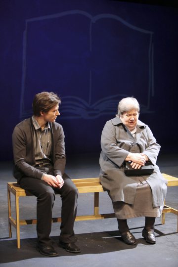 Kaksi ihmistä istuu lavalla puisella penkillä. Vasemmalla istuva nuorempi henkilö katsoo oikealla istuvaa vanhempaa henkilöä, jolla on musta käsilaukku kädessään. Heidän takanaan olevalle siniselle taustalle on piirretty suuri ääriviiva avoimesta kirjasta.