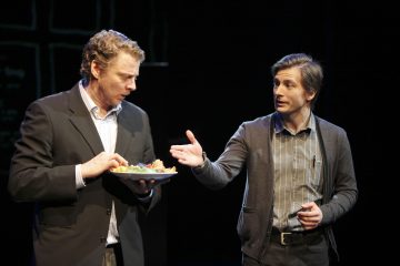 Kaksi miestä lavalla, toinen pukumies, jolla on ruokalautanen kädessään ja hämmentyneen näköinen, kun taas toinen, rennosti pukeutunut, elehtii kädellään kuin selittäisi tai suostuttelisi. Tausta on tumma, mikä viittaa teatteriympäristöön.