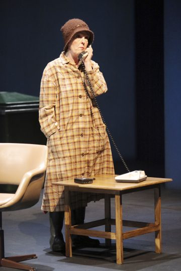 Vanhempi nainen, jolla on ruudullinen takki ja ruskea neulosmyssy, seisoo pienen puupöydän ääressä ja puhuu langalliseen puhelimeen. Lähistöllä hämärästi valaistulla lavalla on beige tuoli ja vihreä roskakori.