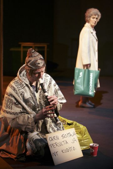 Kuvioituihin vaatteisiin pukeutunut henkilö istuu maassa ja soittaa nauhuria, ja hänen vieressään on käsin kirjoitettu kyltti ja kuppeja. Taustalla katselee vanhempi nainen, jolla on kädessään vihreä ostoskassi.