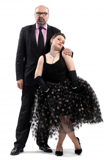 Mustaan pukuun ja vaaleanpunaiseen paitaan pukeutunut mies seisoo käsi mustaan, pilkulliseen tyllimekkoon ja hanskoihin pukeutuneen naisen olkapäällä, joka poseeraa tyylikkäästi miehen edessä valkoista taustaa vasten.