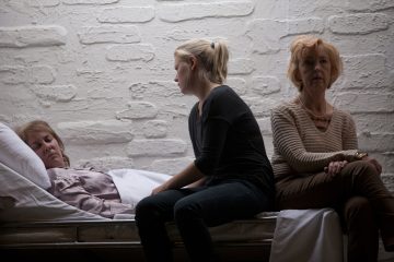 Kolme naista istuu hämärässä huoneessa; yksi heistä makaa sängyssä ja vaikuttaa huonovointiselta, kun taas kaksi muuta istuvat hänen vieressään, synkän ja mietteliään näköisinä valkoista tiiliseinää vasten.