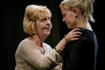 Vanhempi nainen pitelee nuorempaa naista hellästi olkapäistä ja lohduttaa tätä. Nuorempi nainen katsoo alaspäin surullinen ilme kasvoillaan, kun taas vanhempi nainen näyttää puhuvan hänelle ystävällisesti. Molemmat ovat sisätiloissa tummaa taustaa vasten.