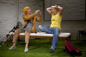 Kaksi ihmistä istuu penkillä; toinen, jolla on päähuivi ja oranssi pusero, näyttää pientä esinettä toiselle, jolla on keltainen paita ja joka näyttää yllättyneeltä, kädet pään päällä. Kainalosauvat ja reppu ovat lähellä.