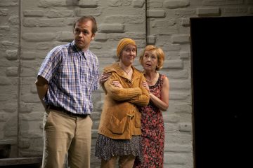 Kolme ihmistä seisoo harmaan tiiliseinän edessä: ruudulliseen paitaan ja khakipukuihin pukeutunut mies katsoo sivulle, kun taas kaksi naista, toinen keltaiseen huiviin ja puseroon pukeutunut ja toinen kukkamekkoon pukeutunut, seisovat lähekkäin ja katsovat eteenpäin.