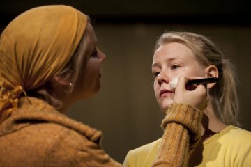 Keltaiseen huiviin ja villapaitaan pukeutunut nainen meikkaa siveltimellä toisen naisen kasvot, jolla on keltainen toppi, pehmeästi valaistussa ympäristössä.