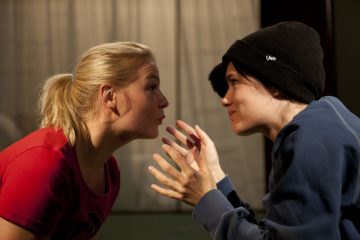 Kaksi ihmistä istuu tiiviisti vastakkain. Toinen, punapaitainen ja vaaleat hiukset taaksepäin sidottu, kuuntelee keskittyneesti. Toinen, jolla on tumma pipo ja sininen collegepaita, elehtii molemmilla käsillään puhuessaan kiihkeästi.