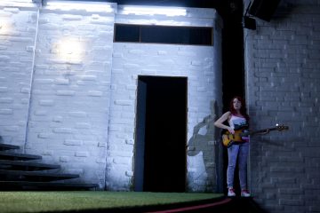 Punatukkainen nainen seisoo oviaukon vieressä pitelemässä sähköbassokitaraa valkoista tiiliseinää vasten kirkkaiden valojen valaisemana. Tila on moderni, vasemmalla on portaikko ja lattialla tekonurmi.