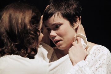 Kaksi valkoisiin vaatteisiin pukeutunutta ihmistä syleilee tiiviisti. Toinen pitelee hellästi toisen kasvoja, joka vaikuttaa tunteelliselta ja kyynelsilmäiseltä ja katsoo kumppaniaan haavoittuvasti. Tausta on tumma, mikä korostaa intiimiä hetkeä.