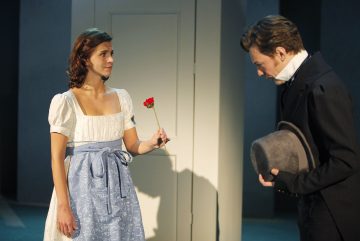 Valkosiniseen ja siniseen mekkoon pukeutunut nainen ojentaa punaisen kukan tummapukuiselle miehelle, jolla on hattu kädessään, kun tämä katsoo alaspäin. He näyttävät olevan näyttämöllä, mikä viittaa teatteriesitykseen.