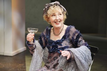 Vanhempi nainen, jolla on kiharat vaaleat hiukset, päällään koristeellinen violetin ja sinisen värinen mekko ja helmiä, hymyilee pitelemässä lasillista samppanjaa, istuu sisätiloissa neutraalia taustaa vasten.