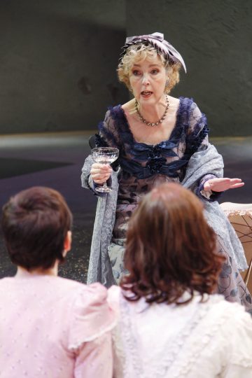 Vanhempi nainen, jolla on koristeellinen sininen mekko ja hattu, pitelee lasia ja elehtii puhuessaan kahdelle nuoremmalle vaaleisiin mekkoihin pukeutuneelle naiselle, jotka istuvat selkä kameraan päin.