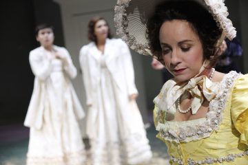 Nainen, jolla on keltainen, 1700-luvun tyylinen mekko ja hattu, seisoo tarkennuksessa ja katsoo alaspäin. Kaksi valkoisiin aikakauden pukuihin pukeutunutta naista näkyy epäselvästi taustalla.