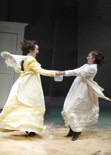 Kaksi naista, joilla on pitkät, koristeelliset aikakauden mekot - toinen keltainen ja toinen valkoinen - pitävät toisiaan kädestä kiinni ja pyörivät iloisesti yhdessä hiekkatäytteisellä lattialla. Heidän hameensa pyörivät, kun he hymyilevät toisilleen teatraalisessa ympäristössä.