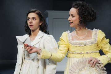 Kaksi aikakauden pukuihin pukeutunutta naista seisoo yhdessä. Vasemmalla oleva, valkoiseen pukeutunut nainen näyttää yllättyneeltä, kun taas oikealla oleva, keltaiseen pukuun pukeutunut nainen hymyilee ja ojentaa hänelle taitetun paperin. Tausta on minimaalinen ja vaimea.