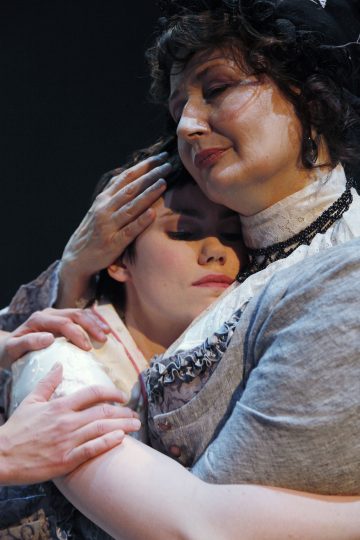 Aikakauden asuun pukeutunut nainen syleilee hellästi toista ihmistä ja laskee päänsä hänen päällensä huolehtiva, suojeleva ilme. Kohtaus viittaa lohdutukseen ja emotionaaliseen tukeen.