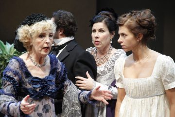 Lavalla esiintyy neljä aikakauden pukuihin pukeutunutta näyttelijää; sinipukuinen vanhempi nainen elehtii eloisasti, kun taas kaksi valkoisiin pukuihin pukeutunutta naista ja pukuinen mies seisovat tarkkaavaisesti, mikä viittaa dramaattiseen tai intensiiviseen kohtaukseen.