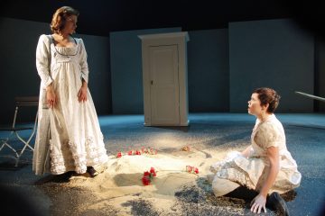 Kaksi valkoisiin aikalaispukuihin pukeutunutta naista esiintyy lavalla. Toinen seisoo tuolin vieressä, toinen polvistuu hiekkaan ja punaisiin kukkiin sirotellun hiekan ja punaisen kukan ääressä, taustalla on valkoinen vaatekaappi ja dramaattinen valaistus.