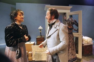 Kaksi aikakauden pukuihin pukeutunutta näyttelijää keskustelee näyttämöllä; toisella on tumma mekko ja hattu, toisella vaalea puku. Taustalla toinen näyttelijä säätää mekkoa vaatekaapin ja vanhojen huonekalujen lähellä.