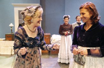 Kaksi aikakauden asuihin pukeutunutta naista hymyilee ja keskustelee keskenään lavasteissa, ja taustalla seisoo kaksi muuta samanlaisiin vintage-asuihin pukeutunutta henkilöä. Kohtaus viittaa vilkkaaseen historialliseen näytelmään tai esitykseen.