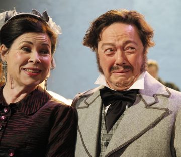 Vintage-asuihin pukeutuneet nainen ja mies seisovat lähekkäin, ja molemmat hymyilevät huvittuneina. Naisella on korvakorut ja tumma mekko, kun taas miehellä on partaharjakset ja suuri rusetti. Tausta on pehmeästi sumea.