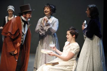 Lavalla on viisi historiallisiin pukuihin pukeutunutta henkilöä. Yksi valkoiseen pukuun pukeutunut istuva henkilö pitelee astiaa ja katsoo hymyillen seisovaa henkilöä, jolla on ruskea viitta ja silinterihattu. Muut seisovat lähellä nauraen ja taputtaen.