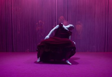 Paljasjalkainen tanssija virtaavassa punaisessa mekossa nojaa dramaattisesti taaksepäin vaaleanpunaisessa ja violetissa valaistuksessa, ja taustalla on pystysuoria jousia ja varjoisia hahmoja, jotka luovat teatraalisen ja tunnelmallisen ilmapiirin.