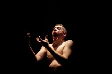 Paidaton henkilö, jolla on dramaattista kasvomaalausta, seisoo pimeässä, suu auki ja kädet kohollaan, valonheittimen valaisemana.